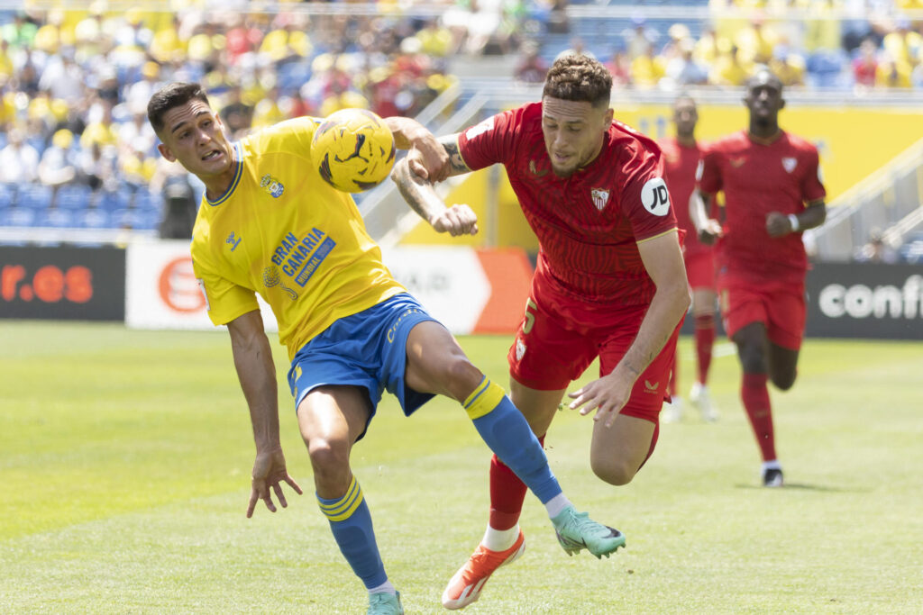 El defensa del Sevilla Lucas Ocampos (d) con el balón ante el defensa del UD Las Palmas Sergi Cardona durante el partido correspondiente a la jornada 31 de LaLiga EA Sports que disputaron en el Estadio de Gran Canaria. EFE/Quique Curbelo